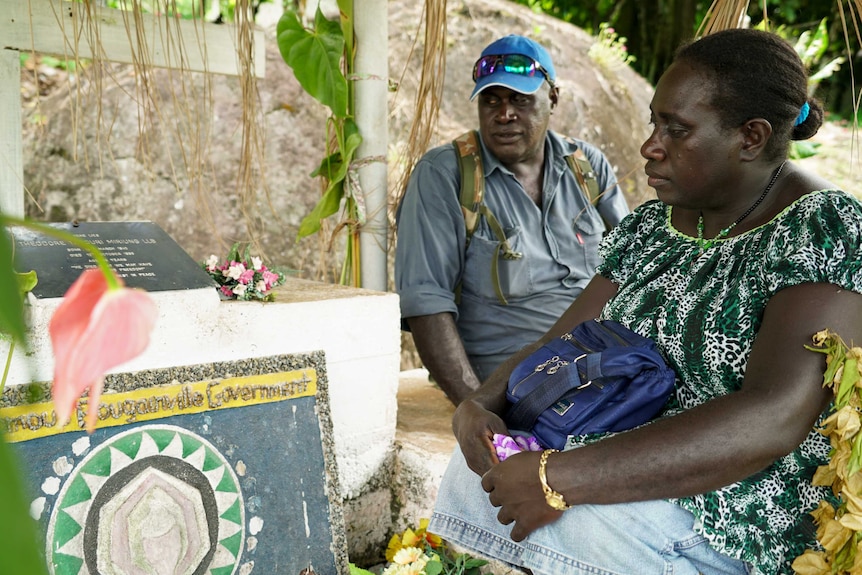 A man and a woman look solemnly at a tombstone