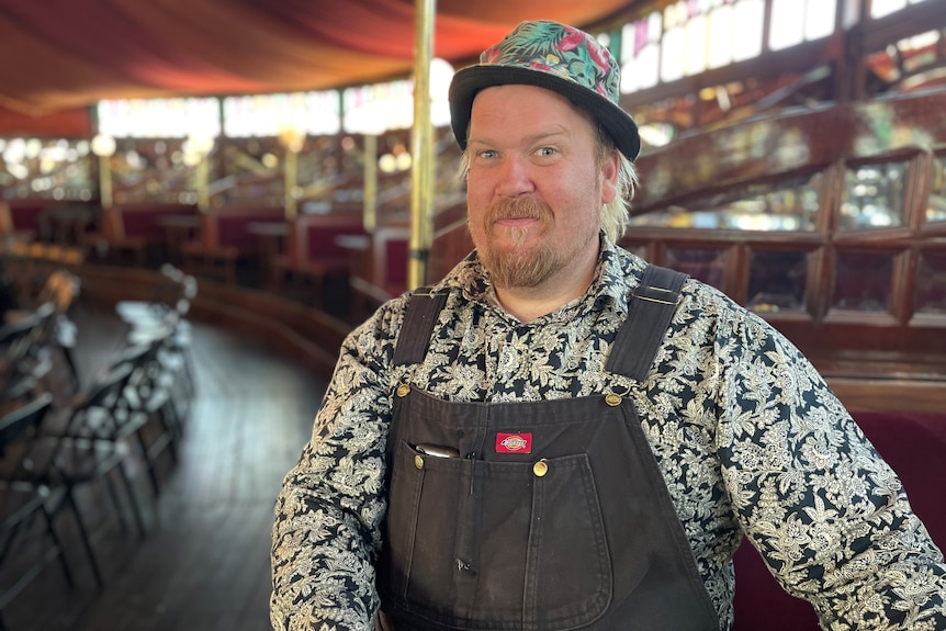 Profile of a man sitting in overalls and a colourful shirt and hat inside a circus tent.