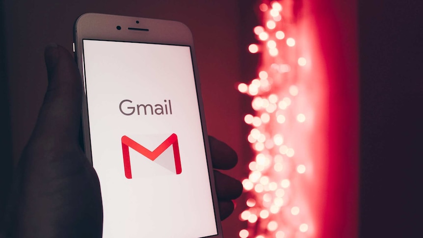 A stock photo of a hand holding a white smartphone loading a Gmail screen with red LEDs in soft focus behind it.