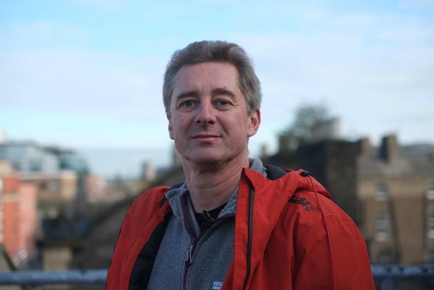 Chris Gray stands outside on a fine day looking at the camera. He is wearing a jumper and jacket.