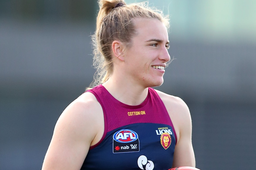 Maria Moloney stands holding an AFL ball wearing a Lions training kit