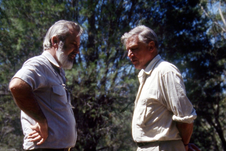 Two men stand together and talk outdoors, they wear shirts.
