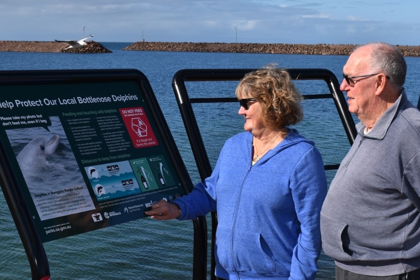 Elderly woman and man on right looking at picture sign in front of ocean marina area