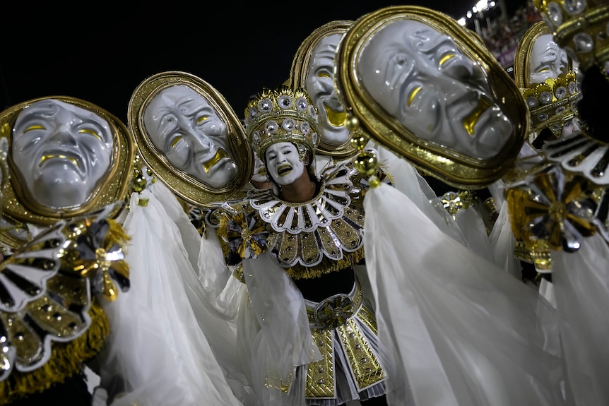 흰색 얼굴 페인트가 있는 흰색과 금색 의상을 입은 댄서가 찡그린 커다란 마스크에 둘러싸여 카메라를 향해 미소를 짓고 있습니다.