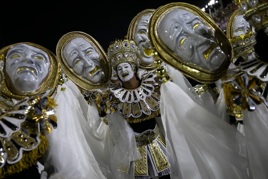 une danseuse vêtue d'une tenue blanche et dorée avec de la peinture blanche pour le visage sourit vers la caméra, entourée de grands masques fronçant les sourcils