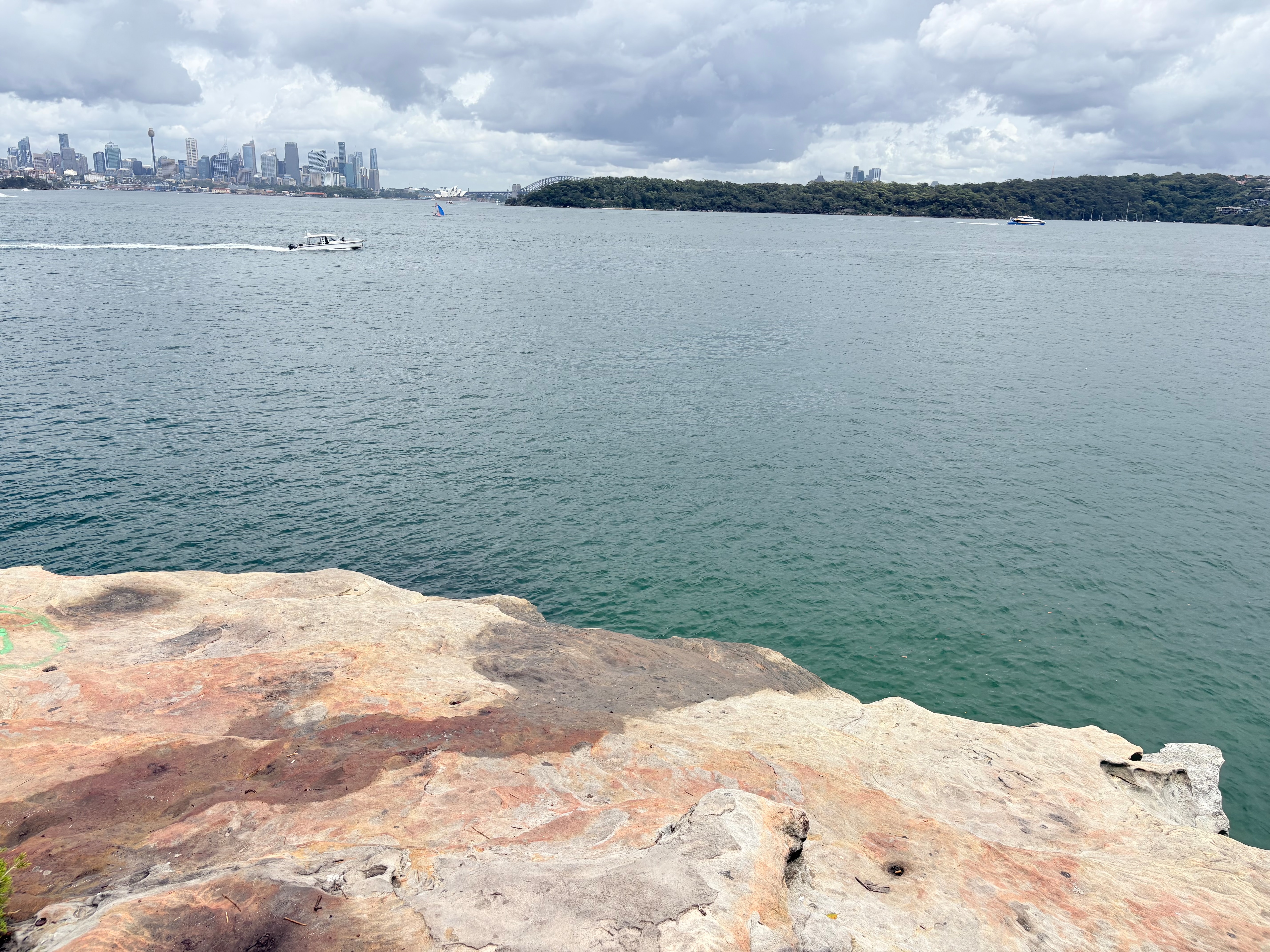 A large flat rock high above the water of Sydney harbour with the bridge and city seen in the distance.