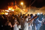 Hundreds of people stand in the street with a building behind them, with smoke billowing above it. Many film something not seen.