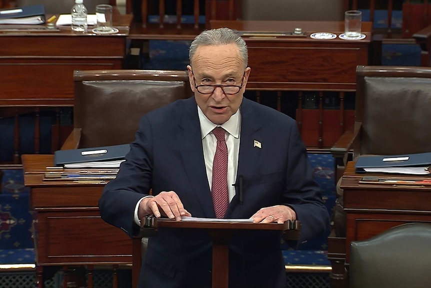 Chuck Schumer wearing a suit, tie and glasses looks up while speaking at a lectern in the US Senate