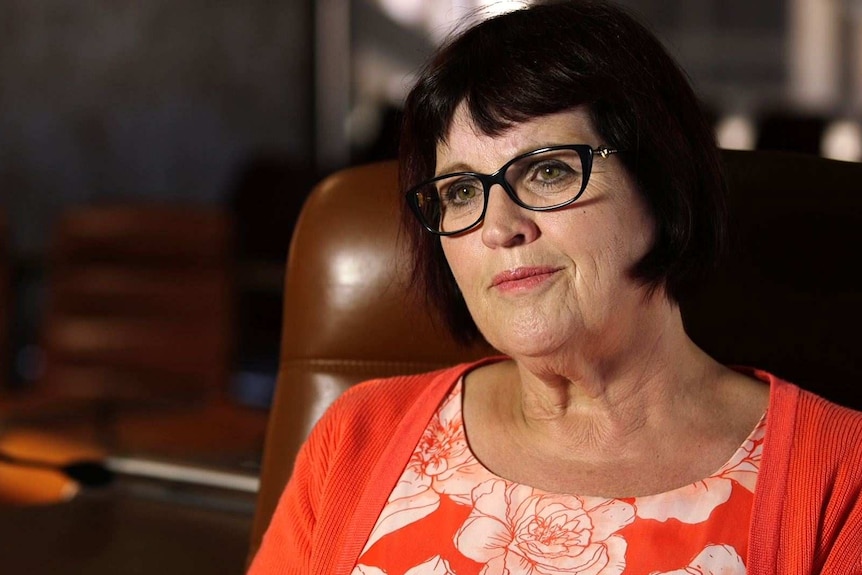 A middle-aged woman with short dark hair wearing glasses and an orange and white top sits on a brown chair.