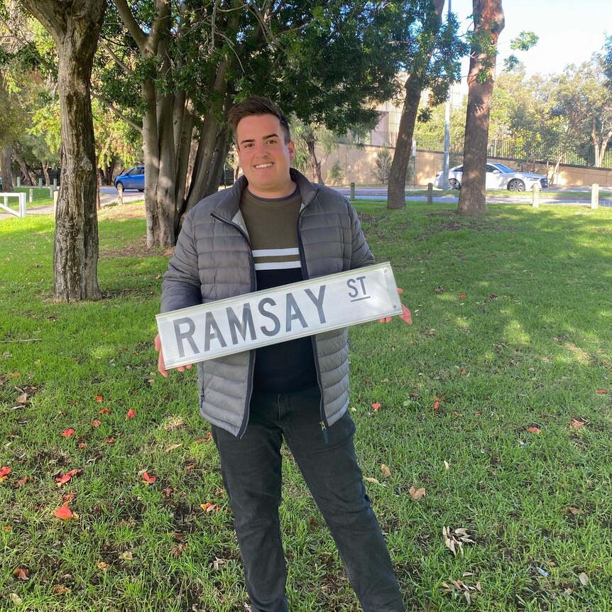 caucasian male with grey puffer jacket holdes ramsay street sign against grassy backdrop