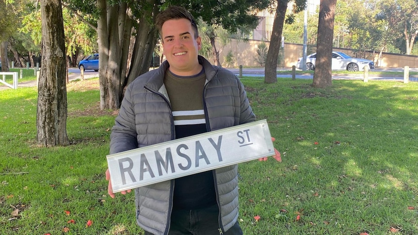 caucasian male with grey puffer jacket holdes ramsay street sign against grassy backdrop