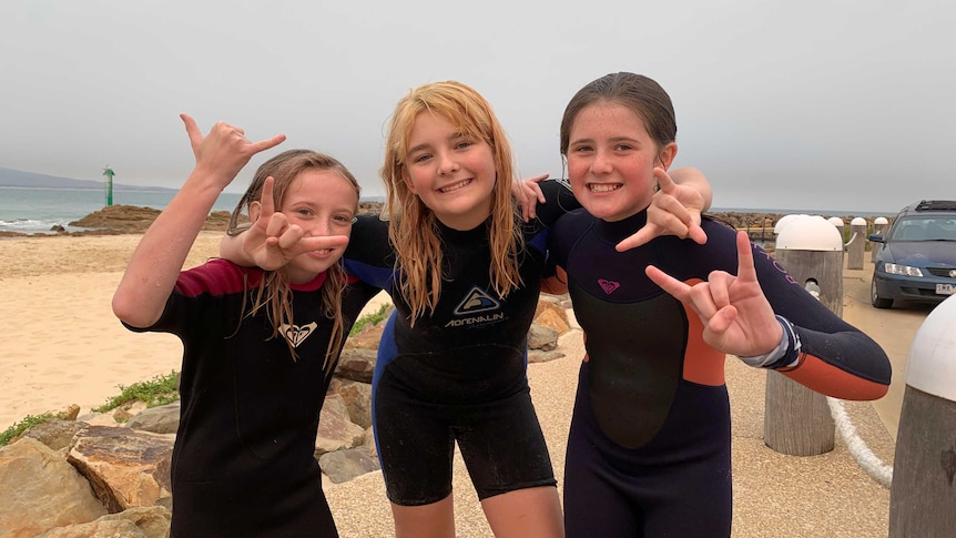Three sisters in wetsuits at the beach. All three are smiling at the camera and pulling the rock n roll sign with their hands.