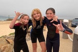 Three sisters in wetsuits at the beach. All three are smiling at the camera and pulling the rock n roll sign with their hands.