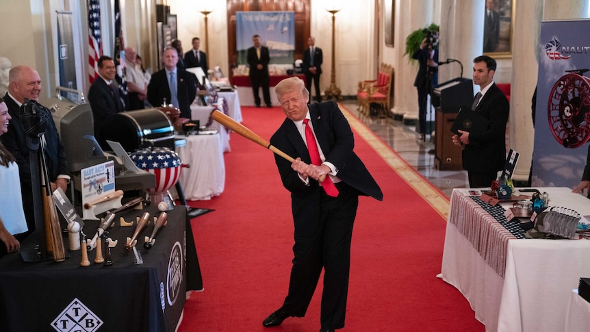 A man with blond hair wearing a suit with a red tie swings a bat in a room while men stand around him.