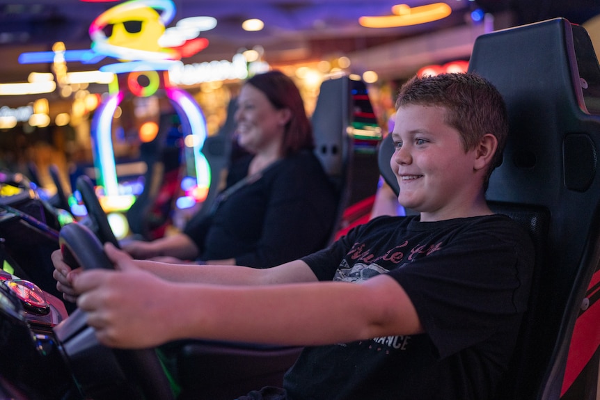 A boy smiles sitting in the driver's seat of an arcade racing game at a neon-lit arcade. His mum smiles in the seat next to him.