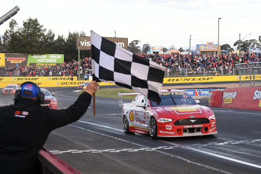A man waves a black and white checquered flag as a racing car speeds over the finish line at Bathurst.