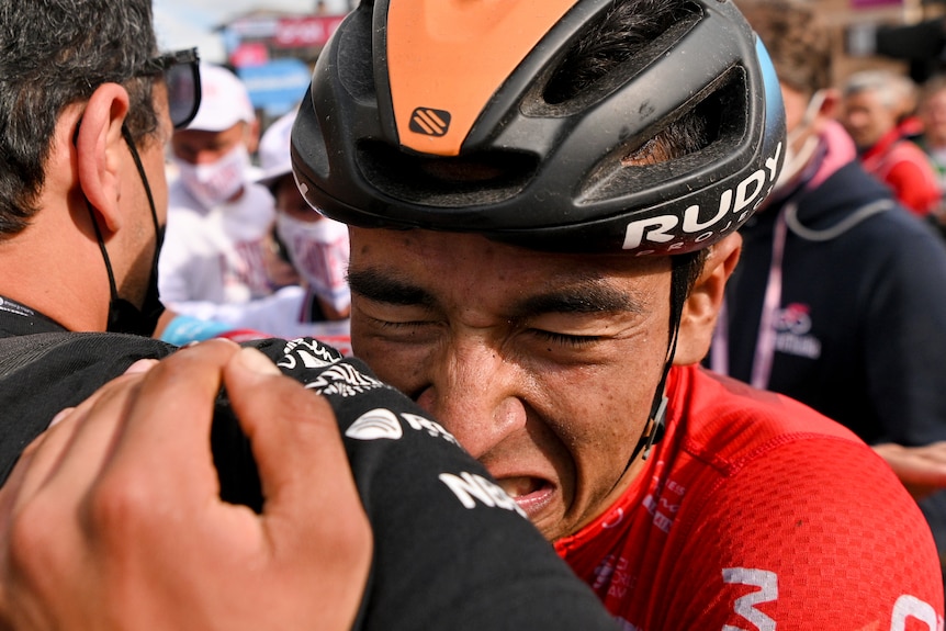 A cyclist hugs a team official after a stage of the Giro d'Italia, his eyes are shut and he is crying 