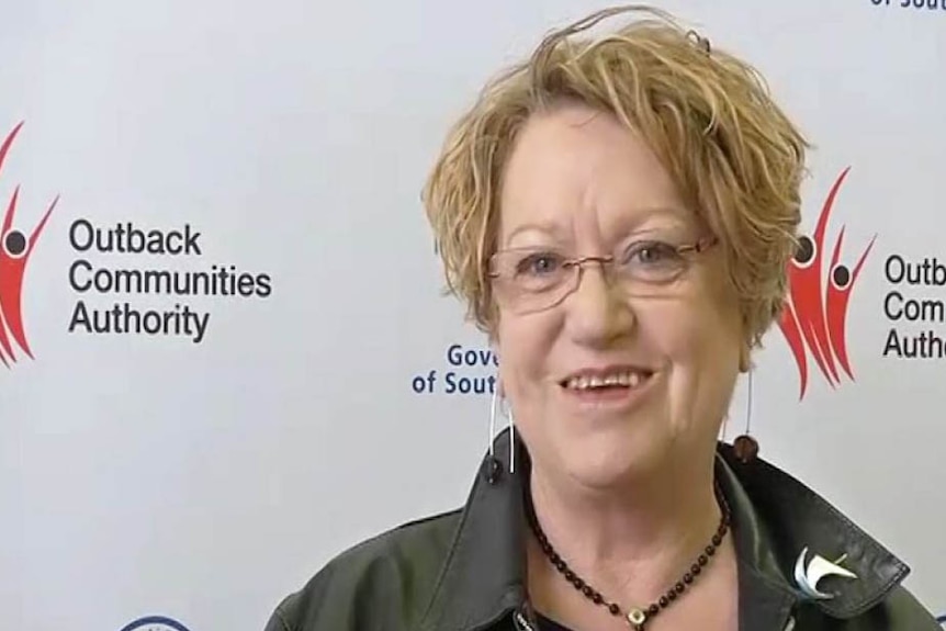 A smiling woman with blonde hair and a leather jacket in front of a screen that reads: "Outback Communities Authority"