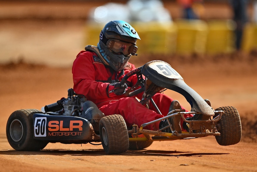 A man in a red jumpsuit racing in a go-kart, on red dirt. One of the front wheels is in the air.