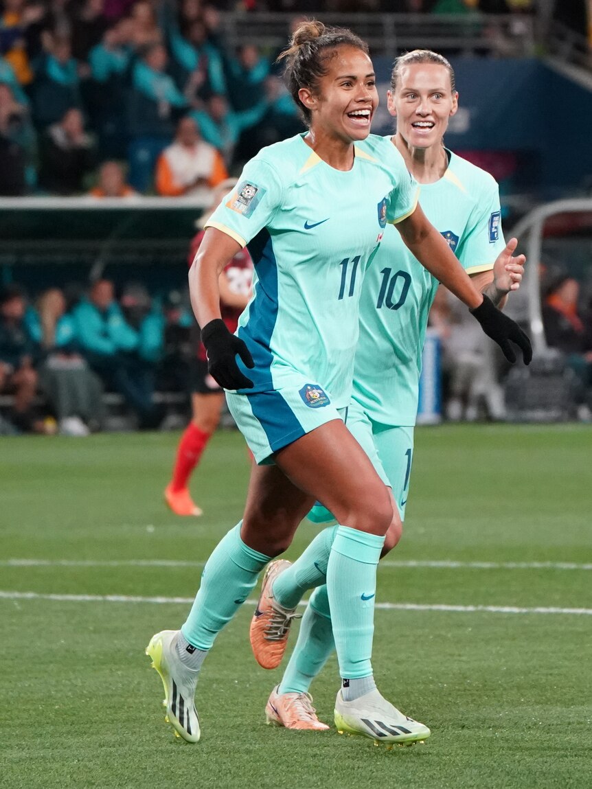 A woman soccer player wearing light blue smiles and runs with her arms out with a teammate behind her