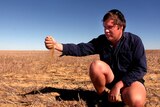 A farmer lifts a handful of dirt amidst dry farmland.