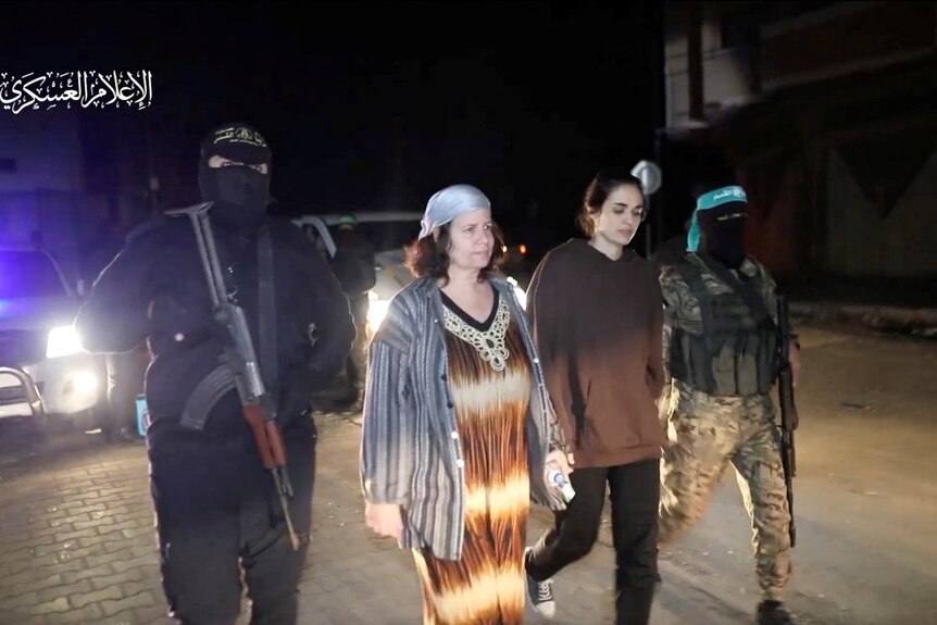 A woman in a brown hoodie walks with a woman in a brown and white dress as they are guided by men with guns.