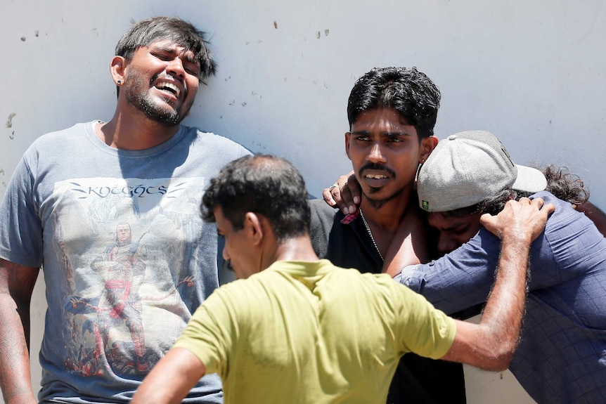 A group of people weep and embrace together against a white brick wall