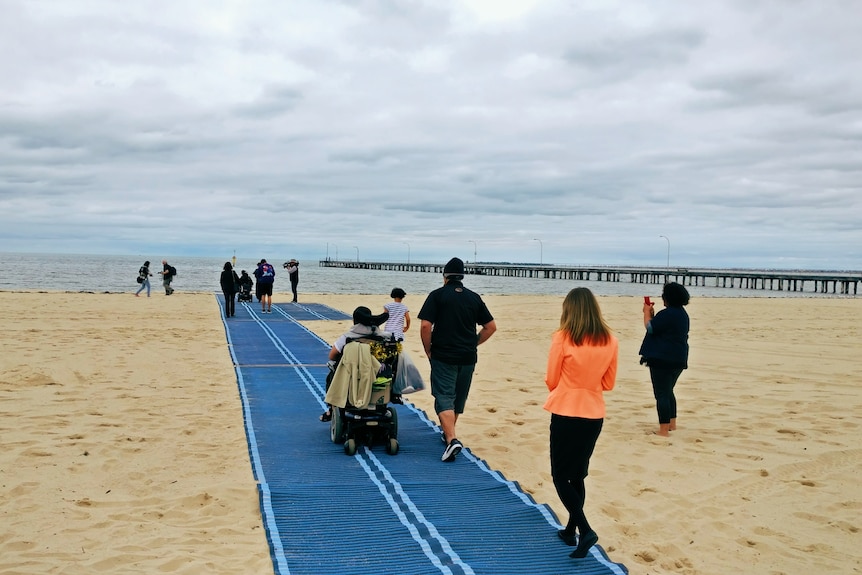 Person on wheelchair is accompanied by others as they travel a blue mat across the beach