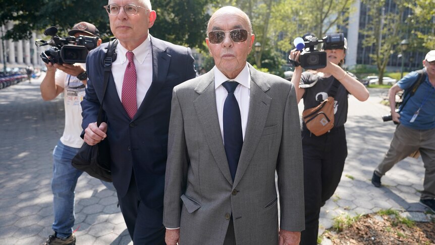 Joe Lewis in a grey suit and aviators walks next to a lawyer as he is followed by camera operators 