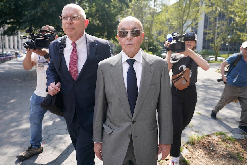 Joe Lewis in a grey suit and aviators walks next to a lawyer as he is followed by camera operators 