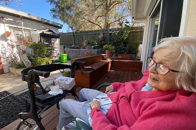 Elderly woman in a bright pink sweater sitting in a backyard.