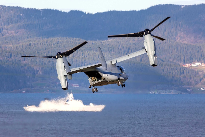 A grey military aircraft with an open rear door flies close to the water