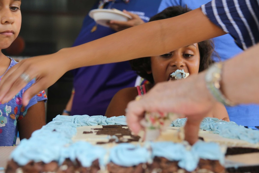 Two Aboriginal children eat pieces cake as an adult hands it out.