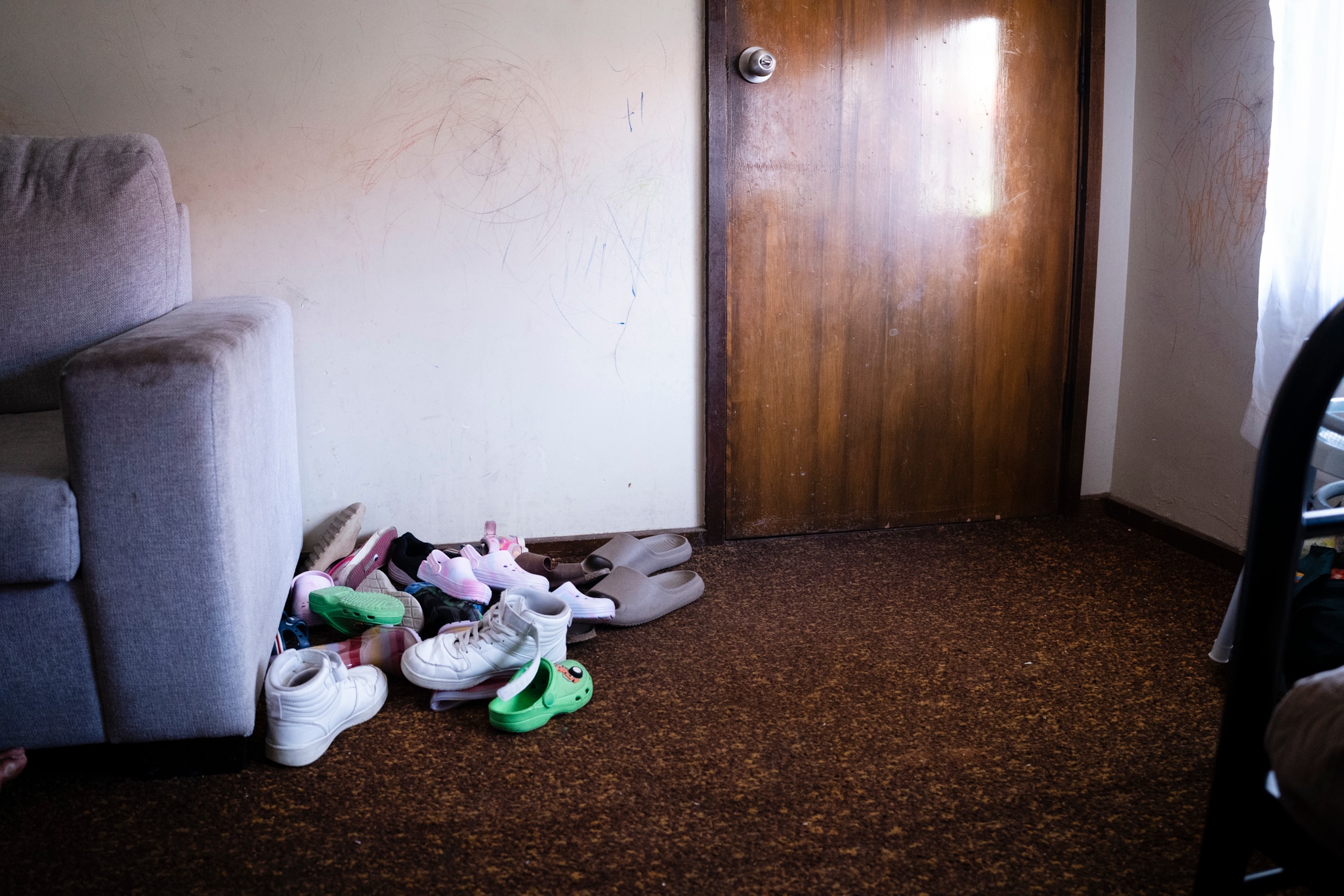 a pile of kids shoes in the corner of a loungeroom with child's drawing on the wall