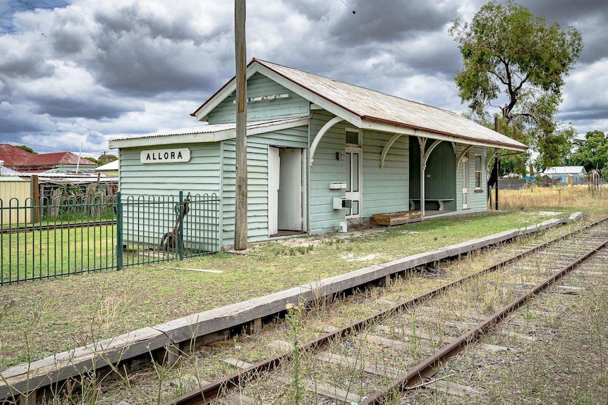 A small turquoise train station next to an overgrown railway line