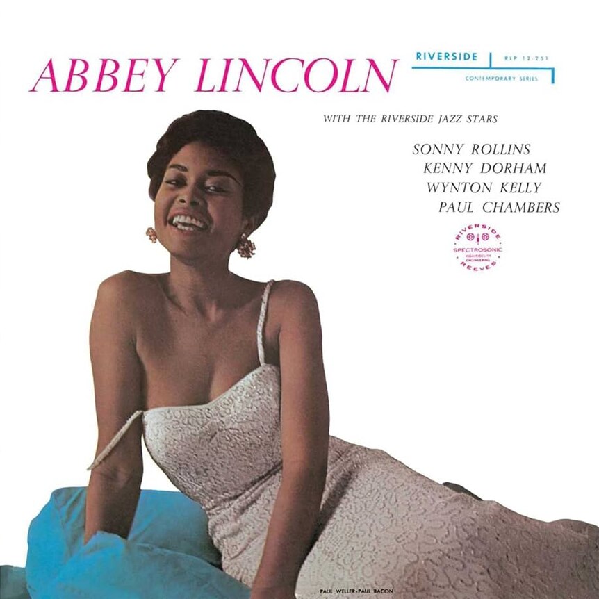 Abbey Lincoln in a silver dress sitting on a blue lounge chair; she's smiling at the camera