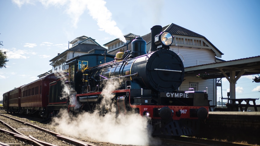 A historic train blows steam on tracks in front of a heritage railway station.
