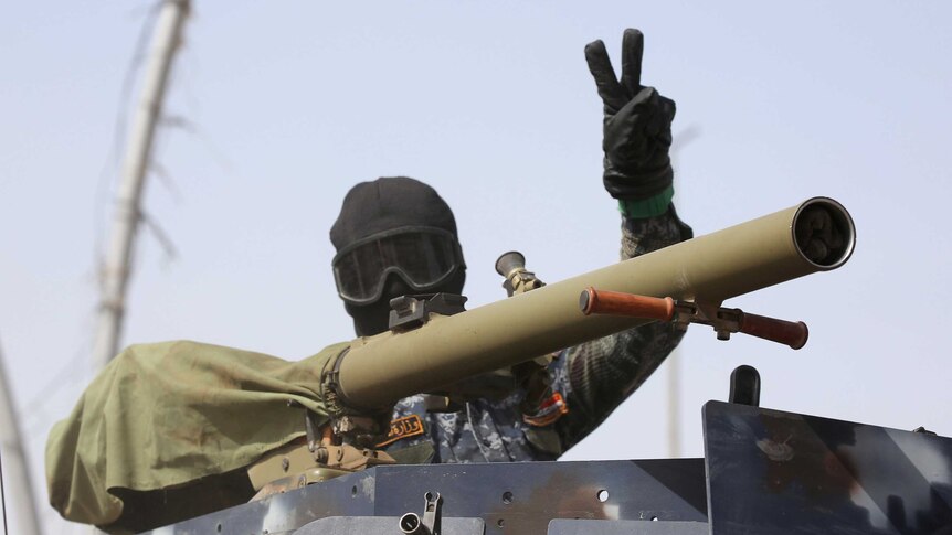 A man whose face is covered with a mask behind a machine gun raises his hand in a peace symbol