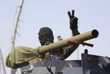 A man whose face is covered with a mask behind a machine gun raises his hand in a peace symbol