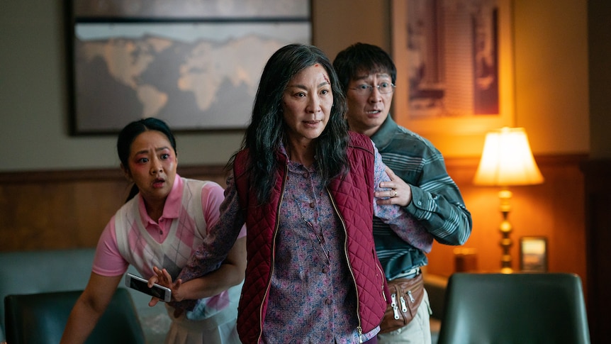 Two Asian-American women and one man in casual attire recoil from something off-camera in a warmly lit hotel room.