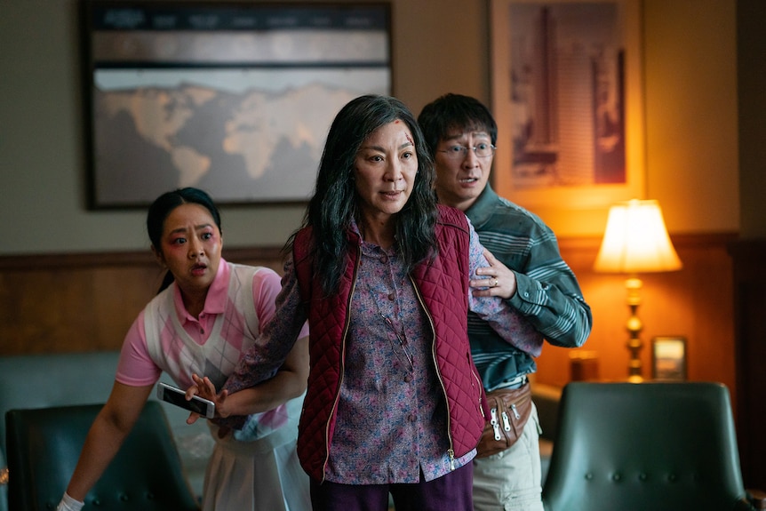 Dos mujeres asiático-estadounidenses y un hombre con ropa informal retroceden ante algo fuera de cámara en una habitación de hotel cálidamente iluminada.