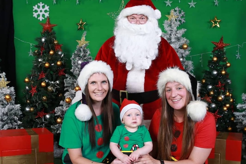 Two women with a baby in green and red and Santa hats with Santa in the background
