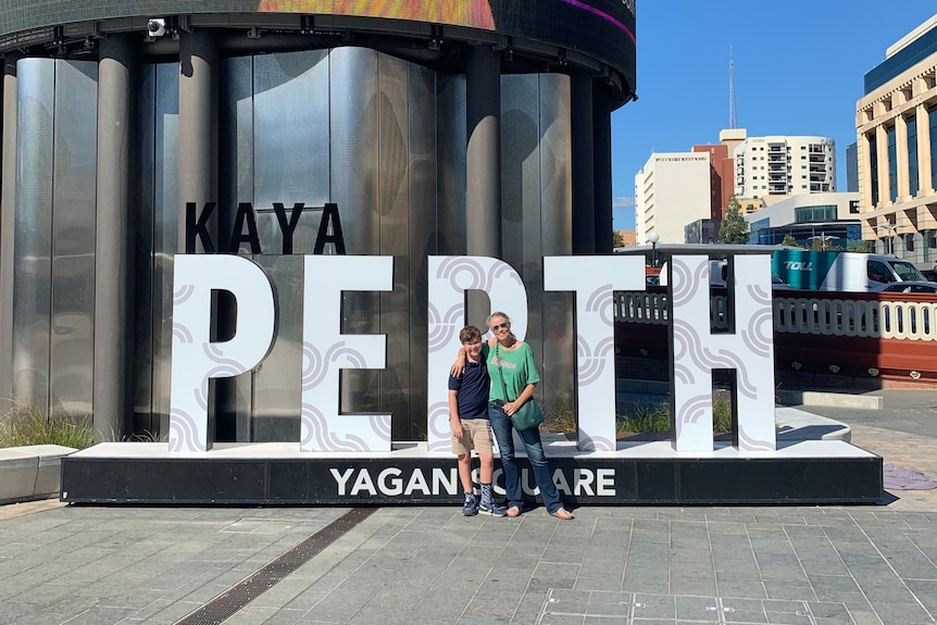 A woman and her son smile happily in front of a sign reading 'PERTH' on a sunny day.