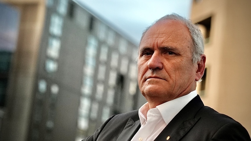 Ken Henry, wearing white open collared shirt and black suit jacket, stands in front of buildings