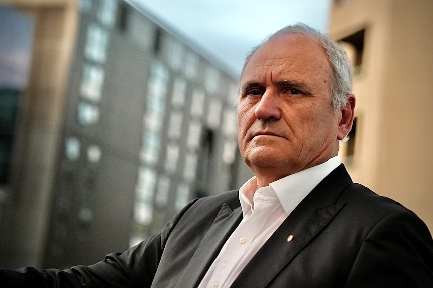 Ken Henry, wearing white open collared shirt and black suit jacket, stands in front of buildings