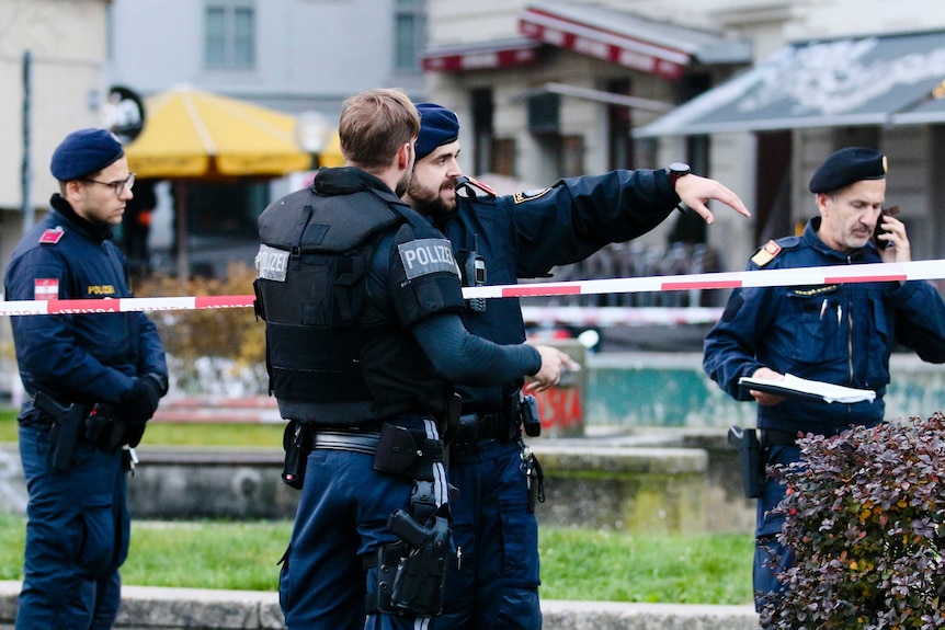 Four armed male police officers in blue stand next to red and white tape in a city square.