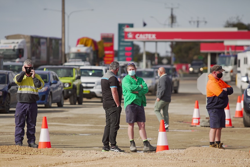 Four men wearing masks are standing in a queue, behind orange and white cones. In the backgound cars are lined up.