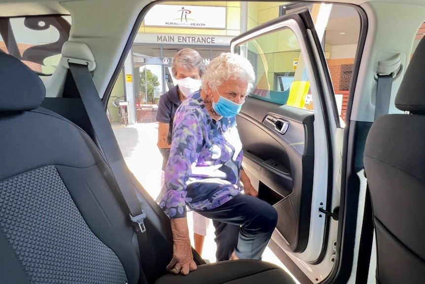 An elderly woman with a blue mask and purple blouse is helped into a car