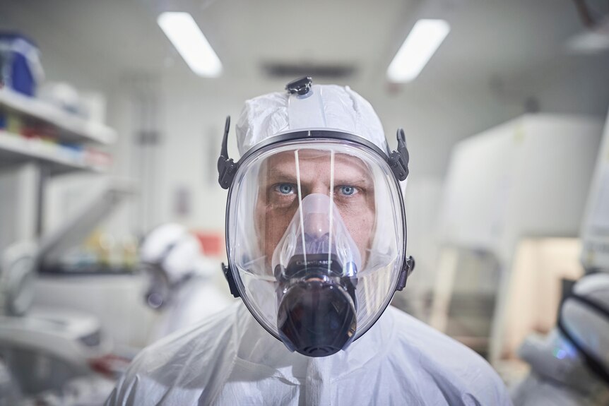 A man wearing a white Tyvek suit and full face PAPR mask with HEPA filter collar poses face on for a photograph in a lab