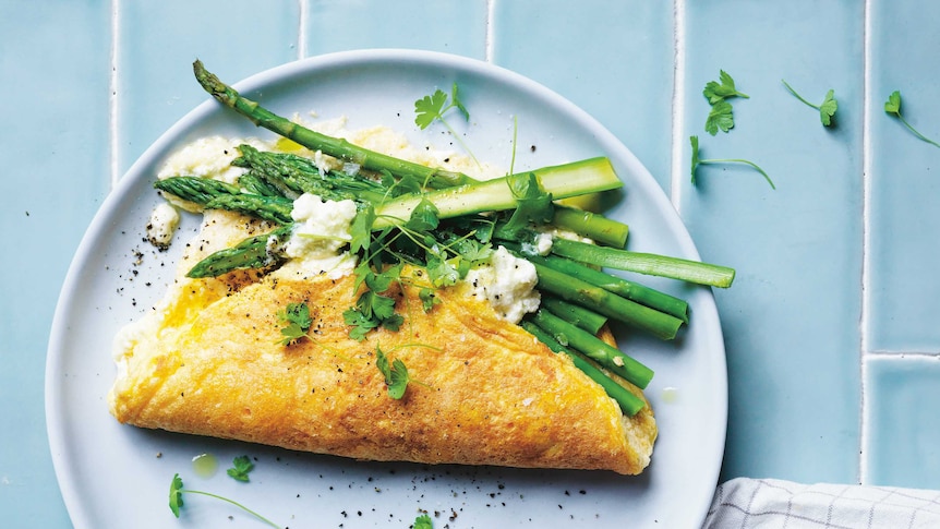 A fluffy folded omelette with asparagus spears dressed in pepper, coriander and feta on a light blue plate set on blue tiles.